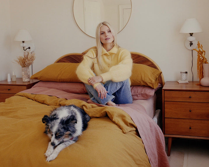 A woman with long blonde hair wearing a yellow sweater and jeans sits cross-legged on a bed with mustard and pink bedding. A small, fluffy black and white dog lies at the foot of the bed. The room is softly lit and decorated in warm tones.