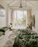 A cozy bedroom featuring Bed Threads’ Olive 100% French Flax Linen Pillowcases, green and beige bedding, a bed by a large window with sheer curtains, a freestanding tub, plants, natural light, and wall-hung towels.