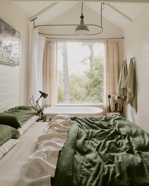 Cozy bedroom with a green and beige bed, a potted plant, and a window overlooking trees and greenery. Light curtains let in natural light, and towels hang by a freestanding bathtub near the window.