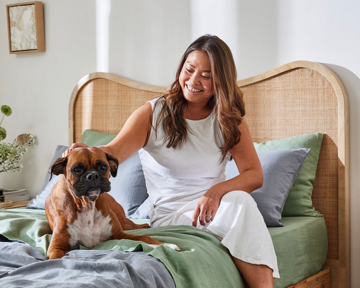 A woman in a white outfit sits on a bed with green and gray bedding, smiling and petting a brown boxer dog beside her. Sunlight fills the cozy, well-decorated bedroom.