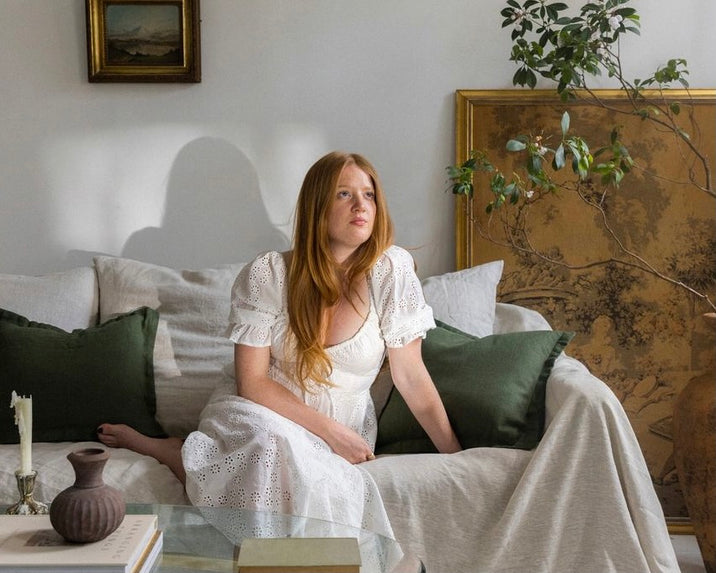 A woman with long red hair in a white dress sits on a light-colored couch with green pillows, surrounded by framed artwork and a potted plant in a softly lit living room.
