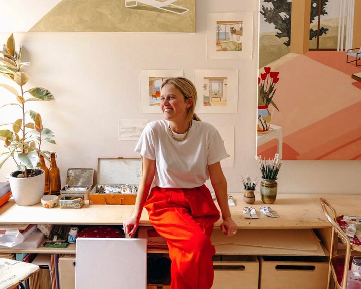 A smiling woman in a white t-shirt and red pants sits on a wooden desk in a bright art studio, surrounded by plants, art supplies, and colorful paintings on the wall behind her.