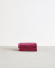 A set of two Berry 100% Cotton Terry Face Washers by Bed Threads, neatly folded in dark pink, is displayed on a light grey surface with a plain white background. - view