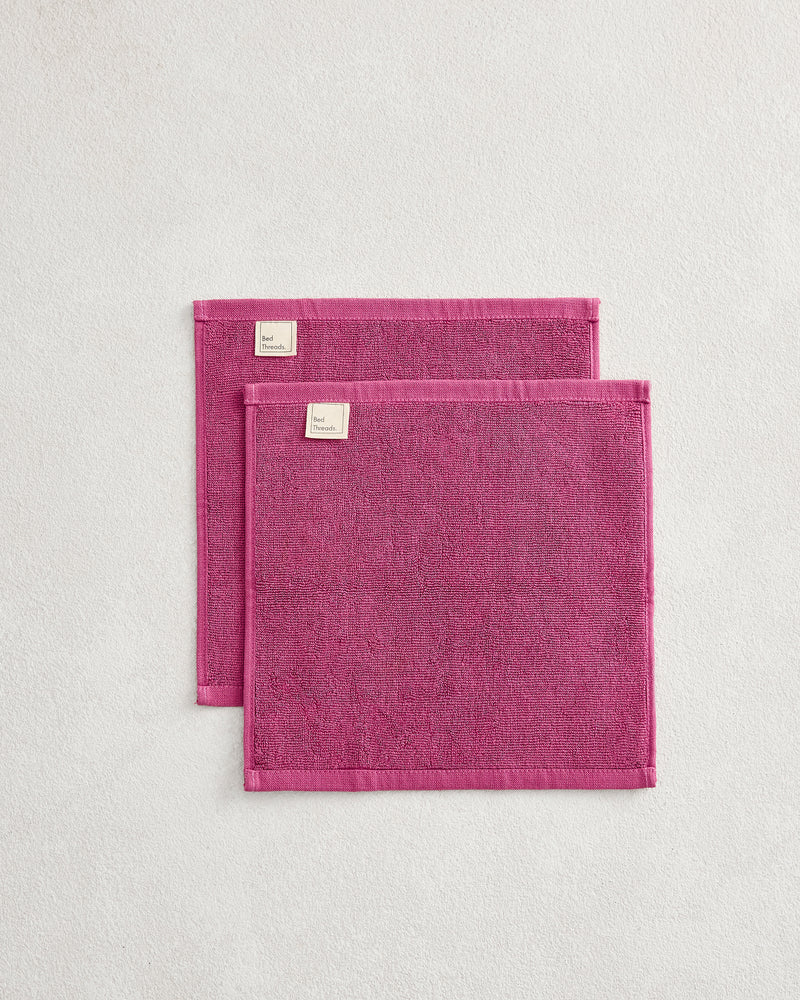 The Berry 100% Cotton Terry Face Washers (Set of Two) by Bed Threads feature soft, absorbent magenta towels with stitched edges and white tags, stacked on a light textured surface—ideal for durable daily face cleansing.