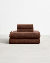 Set of two espresso brown face washers and two hand towels in 100% cotton terry stacked on a light grey surface with a white background.  - view