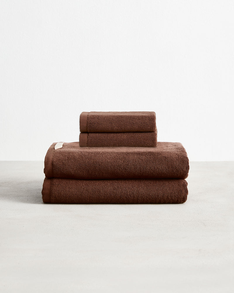 Set of two espresso brown face washers and two hand towels in 100% cotton terry stacked on a light grey surface with a white background. 