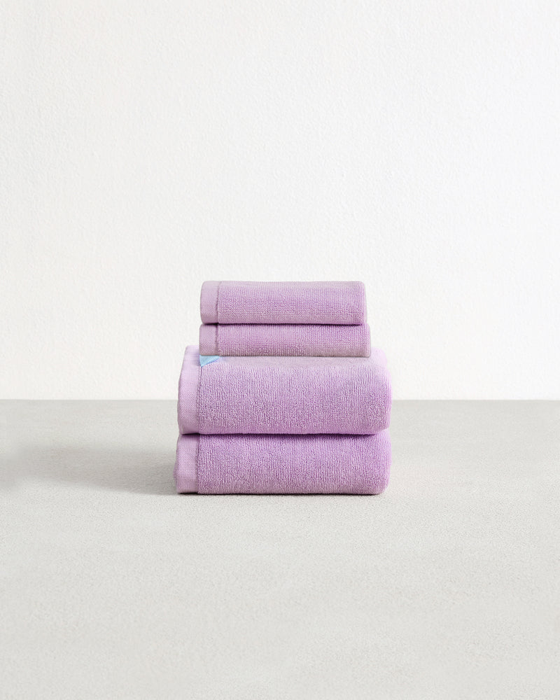 Set of two violet purple face washers and two hand towels in 100% cotton terry stacked on a light grey surface with a white background. 