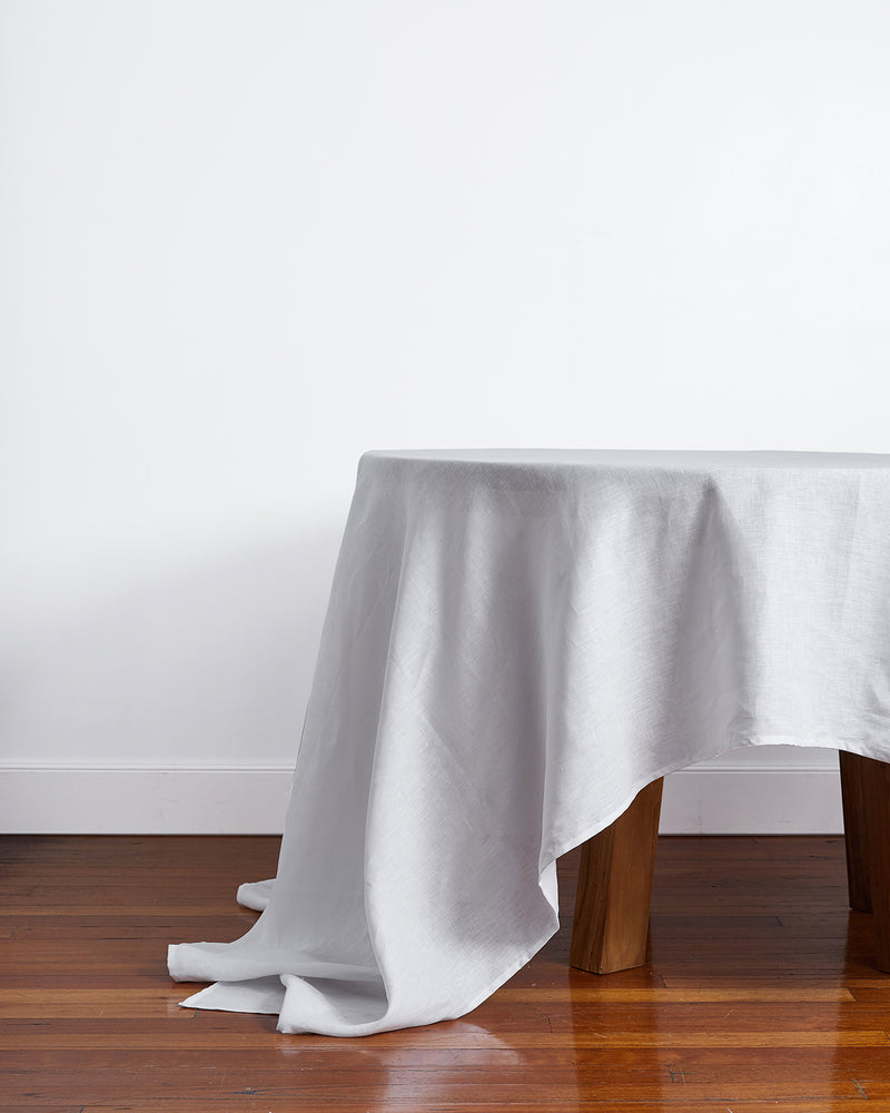 A polished wooden table draped with the Bed Threads White 100% French Flax Linen Tablecloth, its luxurious rectangular fabric pooling onto the floor against a plain white wall.