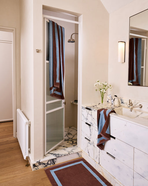 A modern bathroom with a marble sink and shower, featuring Bed Threads’ Cacao & Lagoon Two-Toned 100% Cotton Terry Bath Towels, a matching rug, wood flooring, white flowers in a vase, and a large mirror above the sink.