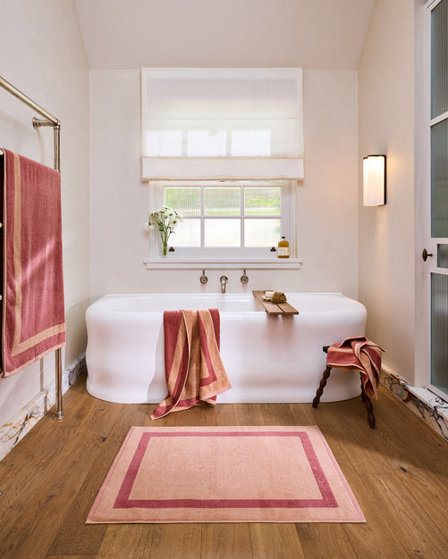 A bright bathroom with a freestanding white tub, Bed Threads Terracotta & Pink Clay Two-Toned 100% Cotton Terry Bath Sheet Bundle on a rack and tub, pink-bordered bath mat, wooden stool, light wood floor, spa accessories, sheer blinds, and wall light.