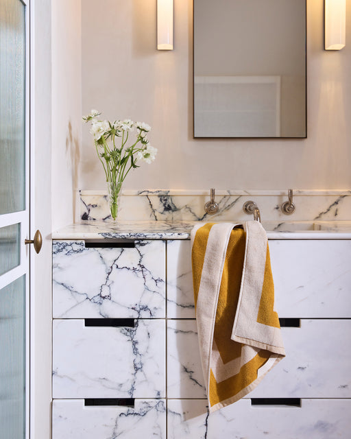 A modern bathroom with a marble vanity, rectangular mirror, wall-mounted faucets, white flowers in a vase, and a Bed Threads Crème & Turmeric Two-Toned 100% Cotton Terry Hand Towel draped over the counter.