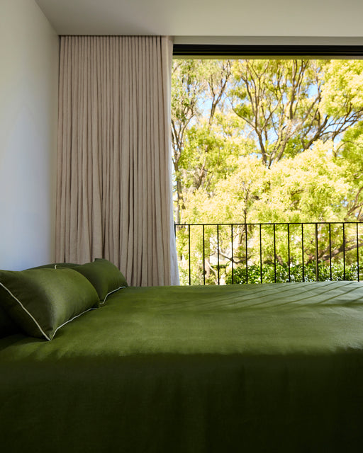 A modern bedroom featuring the Bed Threads Olive Heavy Linen Bed Cover, beige curtains, a large window, and a view of leafy green trees, filling the space with natural sunlight.