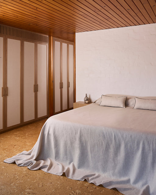 A minimalist bedroom features a bed adorned with Bed Threads’ Oatmeal & Charcoal 100% French Flax Linen Piped Lumbar Cushion Cover, two matching pillows, a white brick wall, wooden closets, a light wood ceiling, and a nightstand with a vase.