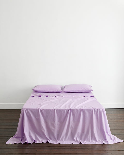 A neatly made bed features Bed Threads Violet 100% Organic Cotton King Pillowcases (Set of Two) against a plain white wall on a dark wooden floor.
