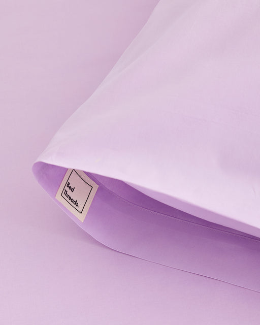 A close-up of Violet 100% Organic Cotton Pillowcases (Set of Two) by Bed Threads on a matching sheet, featuring a small branded tag.