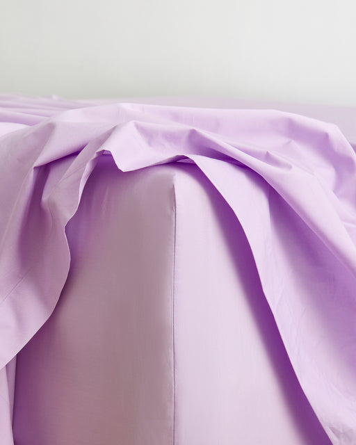 A close-up of a bed corner showcases the Bed Threads Violet 100% Organic Cotton Fitted Sheet with light purple sheets, featuring a loosely draped top sheet for a relaxed, all-natural bedding look.