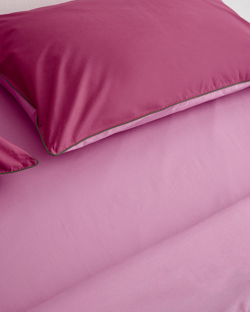 A close-up of a neatly made bed with smooth, pink Bed Threads Berry, Bubblegum & Moss Piped 100% Organic Cotton Pillowcases (Set of Two), showcasing their luxurious, satin-like texture.