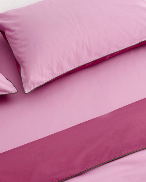Close-up of a bed with Bed Threads’ Berry, Bubblegum & Moss Piped 100% Organic Cotton Pillowcases (Set of Two), showcasing neatly made pink bedding and pillowcases with a contrasting darker pink piped edge.