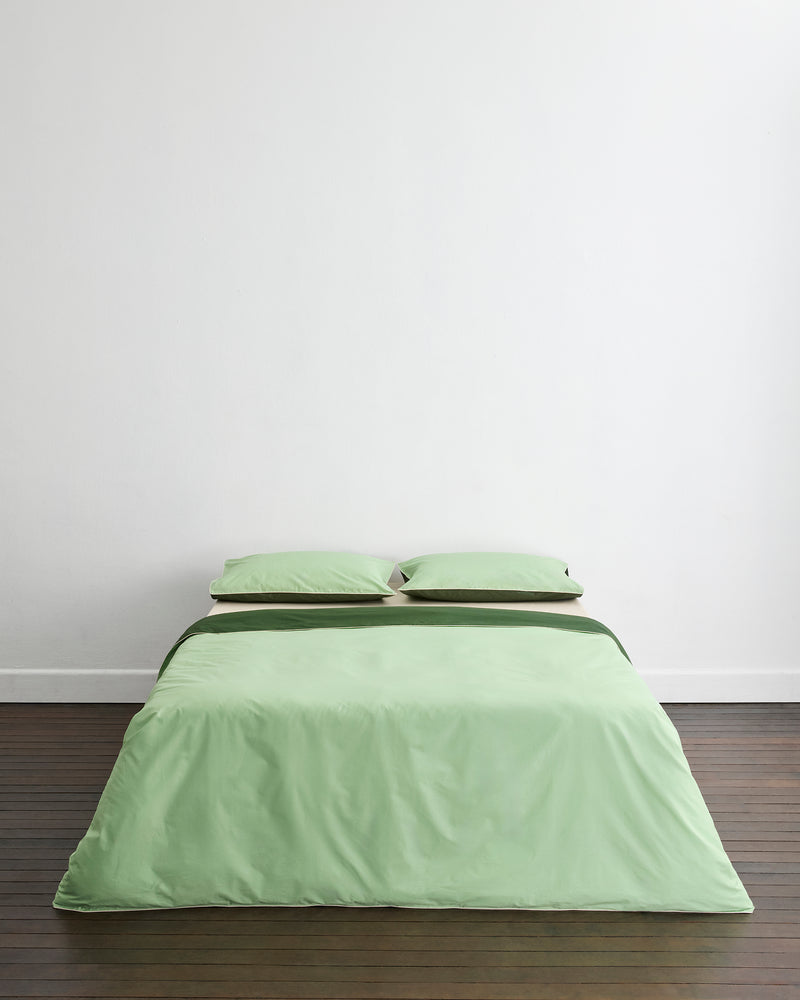 A neatly made bed with light green bedding and Bed Threads’ Moss, Apple & Vanilla Piped 100% Organic Cotton King Pillowcases (Set of Two), set against a plain white wall on a wooden floor.