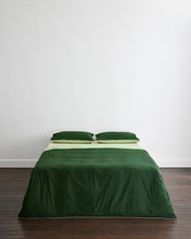 A neatly made bed features Bed Threads' Moss, Apple & Vanilla Piped 100% Organic Cotton King Pillowcases (Set of Two) against a plain white wall on a dark wooden floor. - view