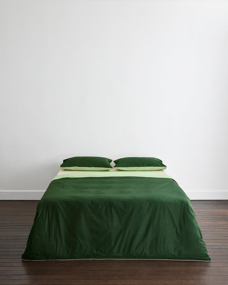A neatly made bed features Bed Threads' Moss, Apple & Vanilla Piped 100% Organic Cotton King Pillowcases (Set of Two) against a plain white wall on a dark wooden floor.