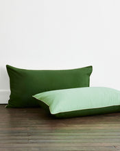 Two pillows featuring Bed Threads’ Moss, Apple & Vanilla Piped 100% Organic Cotton King Pillowcases (Set of Two) rest on a wooden floor—one upright in dark green, the other light green with white piping—for a soft, natural look. - view