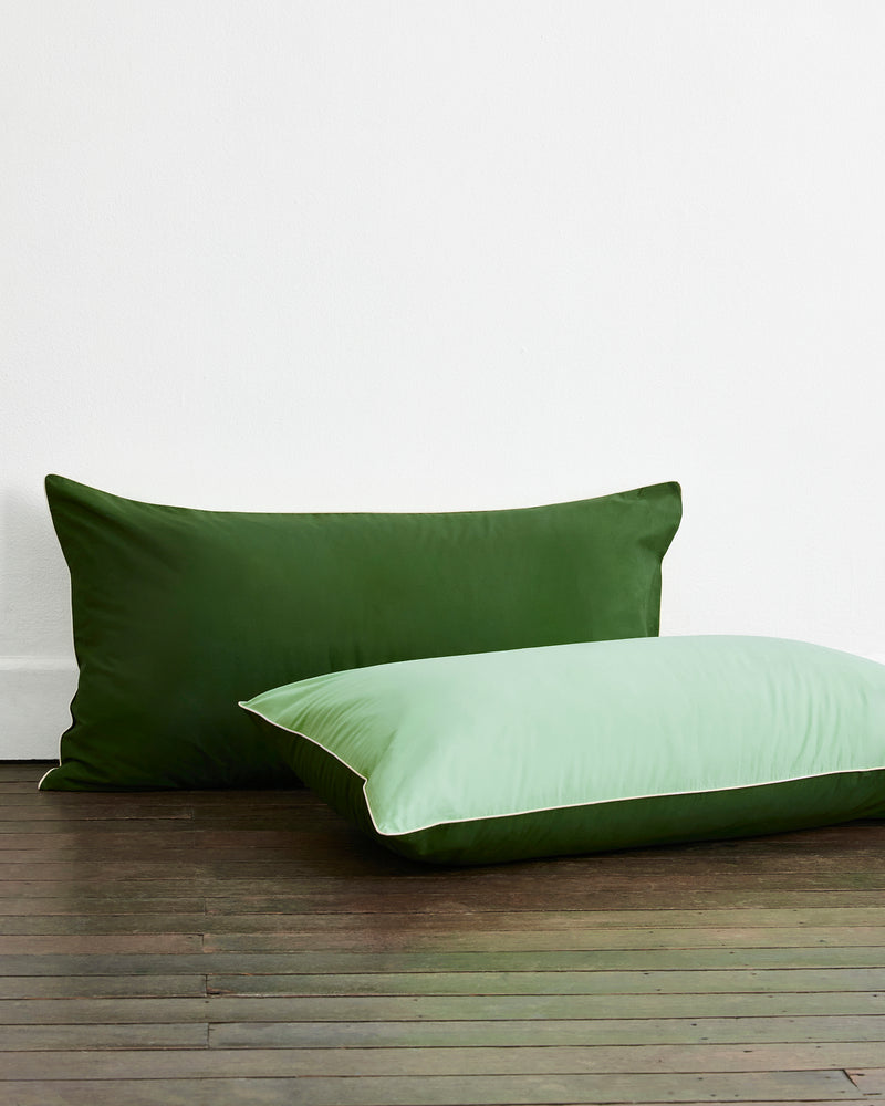 Two pillows featuring Bed Threads’ Moss, Apple & Vanilla Piped 100% Organic Cotton King Pillowcases (Set of Two) rest on a wooden floor—one upright in dark green, the other light green with white piping—for a soft, natural look.