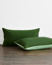 Two Moss, Apple & Vanilla Piped 100% Organic Cotton King Pillowcases by Bed Threads are placed on a wooden floor against a white wall. - view