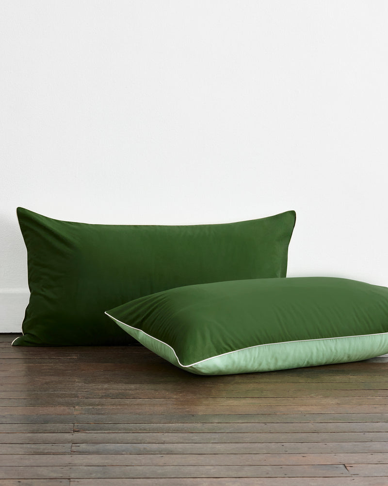 Two Moss, Apple & Vanilla Piped 100% Organic Cotton King Pillowcases by Bed Threads are placed on a wooden floor against a white wall.