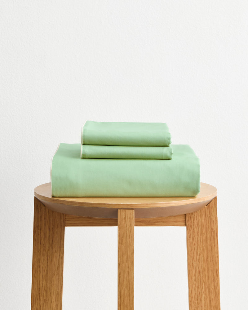 The Bed Threads Moss, Apple & Vanilla Piped 100% Organic Cotton Duvet Cover Set, in light green, is neatly folded and placed on a light wooden stool against a plain white wall background.