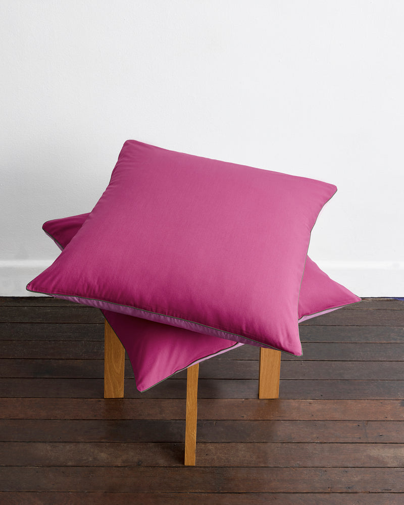 Two Berry, Bubblegum & Moss Piped 100% Organic Cotton European Pillowcases (Set of Two) by Bed Threads are stacked on a four-legged wooden stool, placed on a dark wood floor against a white wall.