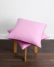 A set of Berry, Bubblegum & Moss Piped 100% Organic Cotton European Pillowcases by Bed Threads is stacked on a simple wooden stool placed on a dark floor against a plain white wall. - view