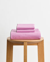 The Berry, Bubblegum & Moss Piped 100% Organic Cotton Duvet Cover Set by Bed Threads, featuring pink GOTS-certified bedding with green trim, is neatly folded on a light wooden stool against a plain white background. - view