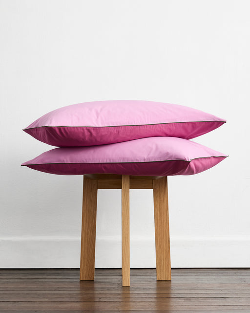 Two Berry, Bubblegum & Moss Piped 100% Organic Cotton Pillowcases (Set of Two) by Bed Threads are stacked on a wooden stool, set against a white wall and wooden floor.