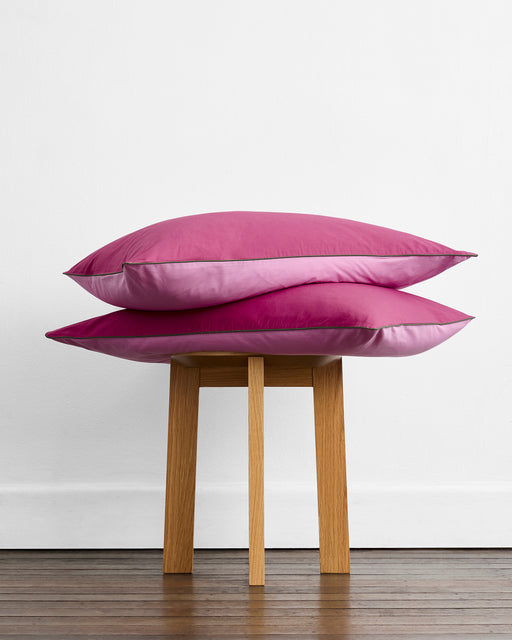 A set of two Bed Threads Berry, Bubblegum & Moss Piped 100% Organic Cotton Pillowcases is stacked on a simple four-legged wooden stool, against a white wall on a wooden floor.