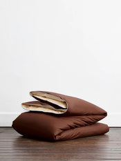 Two neatly folded brown comforters with beige lining rest on a wooden floor against a white wall, perfectly complementing the Bed Threads Espresso, Vanilla & Sky Piped 100% Organic Cotton Bedding Bundle. - view