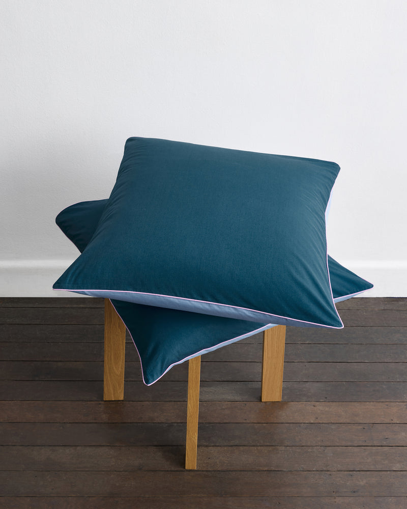 Two Bed Threads Aegean, Sky & Bubblegum Piped 100% Organic Cotton European Pillowcases (set of two) are stacked on a small wooden stool, atop a dark wood floor in front of a plain white wall.