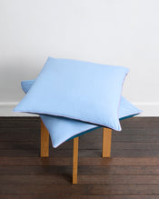 Two Aegean, Sky & Bubblegum Piped 100% Organic Cotton European Pillowcases by Bed Threads are stacked on a small wooden table with a dark wood floor and plain white wall in the background. Set includes two pillowcases. - view
