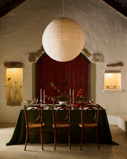 A dining table set for four with a green tablecloth is styled with Bed Threads’ Mulberry 100% French Flax Linen Napkins (Set of Four), beneath a round paper lantern in a rustic room featuring a red arched door, stone walls, and niches with plants and candles.