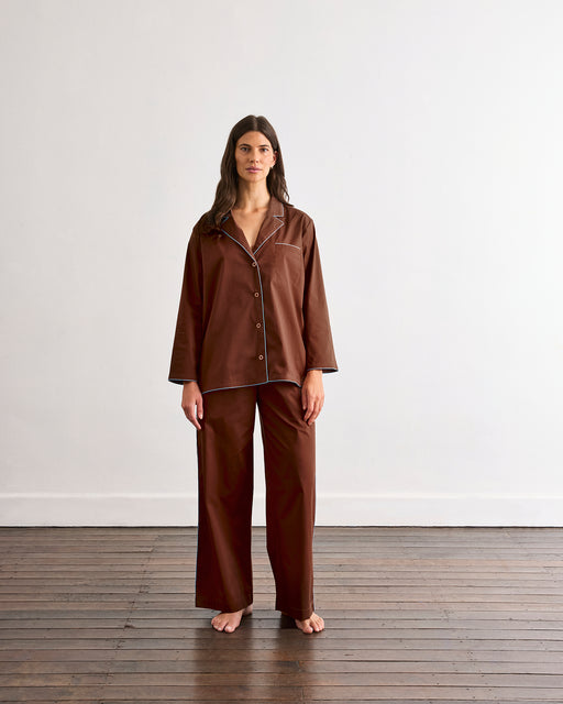 A woman stands barefoot on a wooden floor in front of a plain white wall, wearing the Bed Threads Espresso & Sky Piped Long Sleep Set.