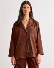 A woman with long brown hair faces the camera, wearing the Bed Threads Espresso & Sky Piped Long Sleep Set—loose-fitting with white piping and matching pants—against a plain light background. - view