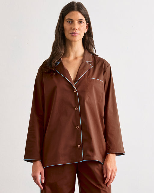 A woman with long brown hair faces the camera, wearing the Bed Threads Espresso & Sky Piped Long Sleep Set—loose-fitting with white piping and matching pants—against a plain light background.