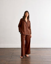A woman stands barefoot on a wooden floor in the Bed Threads Espresso & Sky Piped Long Sleeve Shirt, a brown, loose-fitting cotton top with subtle contrast piping, set against a plain white wall. - view
