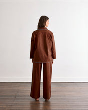 A woman faces away from the camera, wearing the Bed Threads Espresso & Sky Piped Long Sleeve Shirt—a brown cotton pyjama top with light blue piping—paired with matching wide-leg pants. - view
