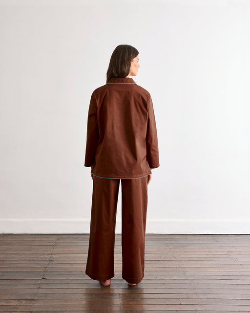 A woman faces away from the camera, wearing the Bed Threads Espresso & Sky Piped Long Sleeve Shirt—a brown cotton pyjama top with light blue piping—paired with matching wide-leg pants.