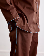 A person wearing Bed Threads' Espresso & Sky Piped Pants, shown from shoulders to knees, features brown relaxed-fit pants with light blue piping and an elasticated waist, one hand in their pocket. - view