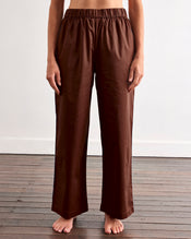 A person stands barefoot on a wooden floor, wearing Bed Threads' Espresso & Sky Piped Pants—relaxed, breathable cotton with an elastic waist and side pockets—against a plain white wall. - view