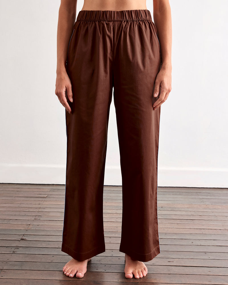 A person stands barefoot on a wooden floor, wearing Bed Threads' Espresso & Sky Piped Pants—relaxed, breathable cotton with an elastic waist and side pockets—against a plain white wall.