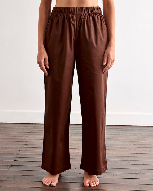 A person stands barefoot on a wooden floor, wearing the Bed Threads Espresso & Sky Piped Long Sleep Set—loose-fitting pants with an elastic waistband and side pockets—against a plain white wall.