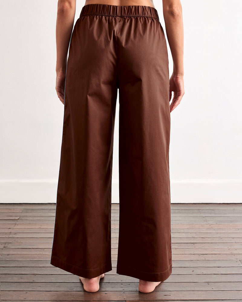 A person stands barefoot on wooden floorboards, seen from behind, wearing Bed Threads' Espresso & Sky Piped Pants with an elastic waist in breathable cotton. A plain white wall forms the background.